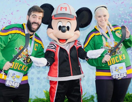 Katie and Spencer wearing Mighty Ducks jerseys carrying hockey sticks with Mickey Mouse wearing sports outfit after 2020 Walt Disney World 10K